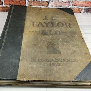 May include: An antique book with a worn, tan cover and a black spine. The cover features the text "J.L. TAYLOR & CO." and "SPRING & SUMMER 1917." The book is resting on a white wooden surface.