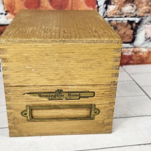 May include: A light brown wooden index card box with finger joint corners. The front features a black logo with the text Remington Rand Made in U.S.A. and a brass metal label holder attached with two small screws centered below the logo.