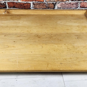 May include: A rectangular wooden tray with raised edges. The tray is made of light-colored wood and has a natural finish. The tray is designed to hold items and is suitable for serving or display.