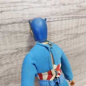 May include: A blue and red action figure with a blue helmet and a red, white, and blue striped shirt. The figure is wearing a blue belt and has a button on the back.