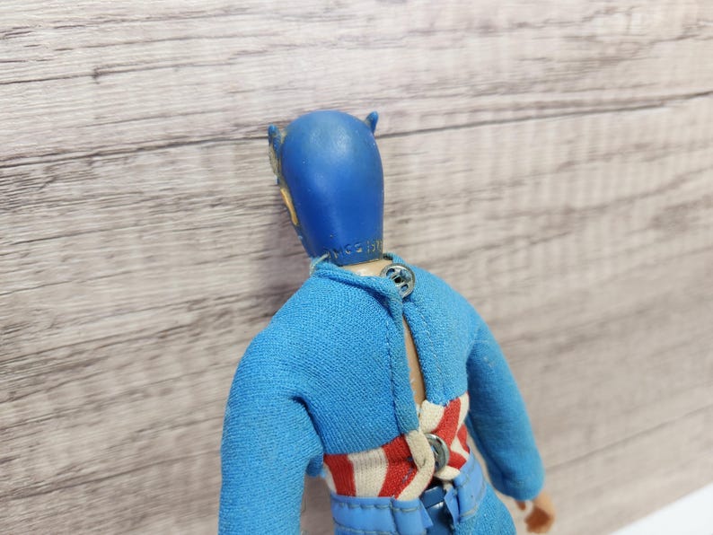 May include: A blue and red action figure with a blue helmet and a red, white, and blue striped shirt. The figure is wearing a blue belt and has a button on the back.