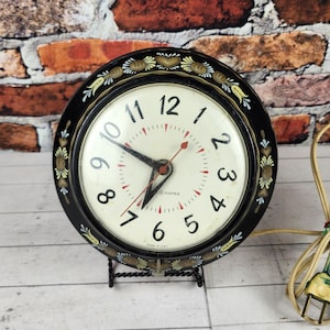 May include: A vintage black and gold clock with a cream-colored face and red second hand. The clock has a decorative floral border and sits on a black metal stand. A tan power cord is bundled to the side.