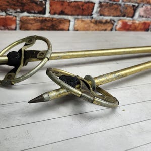May include: Two vintage ski poles with gold-colored shafts and metal rings. The poles have pointed tips and black hand grips. The background features a brick wall and a wooden surface.