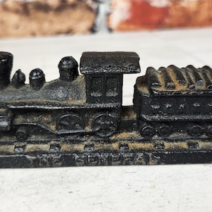 May include: A small, antique-style black train figurine. The locomotive and attached car are detailed with wheels, smokestacks, and a roof. The train sits on a track base, suggesting a vintage or collectible item.