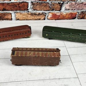 May include: Three vintage toy train cars in shades of brown and green are arranged on a white wooden surface. The cars are rectangular with detailed features, including windows and text. The background features a brick wall.