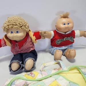 May include: Two Cabbage Patch Kids dolls, one with blonde hair wearing a red and white checkered shirt and blue jeans, and the other with blonde hair wearing a red shirt with the Cabbage Patch Kids logo and blue and white shorts. Both dolls are sitting on a yellow and green patchwork blanket.