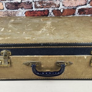 May include: A rectangular vintage suitcase featuring a beige textured exterior with a dark blue trim along the center and edges. The luggage includes a dark blue plastic handle, two metallic latches with keyholes, and visible surface wear.