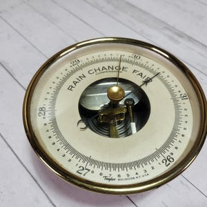 May include: A vintage barometer with a brass-colored frame and a cream-colored face. The face displays the words "Rain Change Fair" and numerical pressure readings. The barometer is a Taylor Rochester NY USA model.
