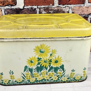 May include: Vintage rectangular tin with a yellow lid and floral design. The off-white body features yellow sunflowers and green leaves. The tin is likely used for storage and has a slightly distressed appearance.