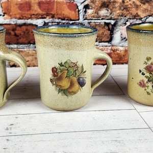 May include: Three ceramic mugs with a beige and tan speckled finish. Each mug features a different fruit or floral design. The mugs have a blue rim and a curved handle. The mugs are displayed on a white wooden surface.