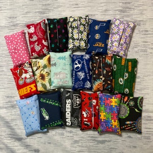 May include: A collection of 18 colorful fabric tissue box covers. The covers feature various designs, including sports teams, animals, and abstract patterns. The covers are all rectangular and have a soft, quilted texture.