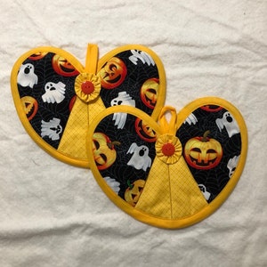 May include: Two heart-shaped pot holders featuring a black fabric background with a white spiderweb pattern, orange jack-o'-lanterns, and white ghosts. Each pot holder has a yellow fabric accent, a yellow fabric loop, and a small red button.