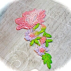 May include: A delicate floral lace applique featuring a large pink rose and smaller pink and purple flowers with green leaves.