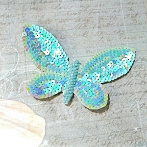 May include: A turquoise blue butterfly-shaped sequin applique. The sequins are iridescent and have a light blue, green, and white color scheme.