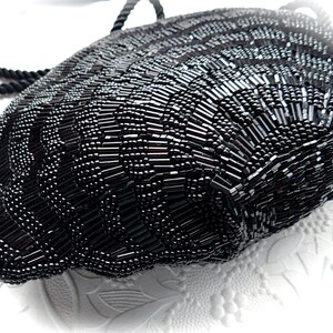Puede incluir: Un bolso de noche negro de cuentas con asa de cuerda. El bolso est&aacute; hecho de peque&ntilde;as cuentas negras que est&aacute;n cosidas juntas en un patr&oacute;n. El bolso tiene forma de semic&iacute;rculo y tiene un fondo redondeado.