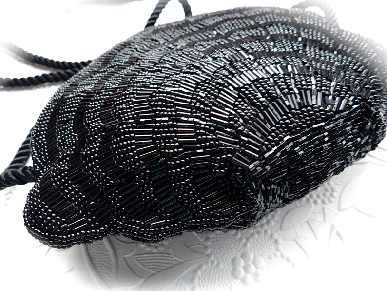 Puede incluir: Un bolso de noche negro de cuentas con asa de cuerda. El bolso est&aacute; hecho de peque&ntilde;as cuentas negras que est&aacute;n cosidas juntas en un patr&oacute;n. El bolso tiene forma de semic&iacute;rculo y tiene un fondo redondeado.