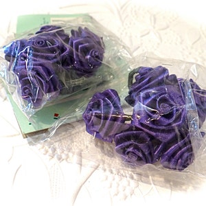 May include: Two packs of purple satin ribbon roses. The roses are arranged in a cluster and are wrapped in clear plastic.
