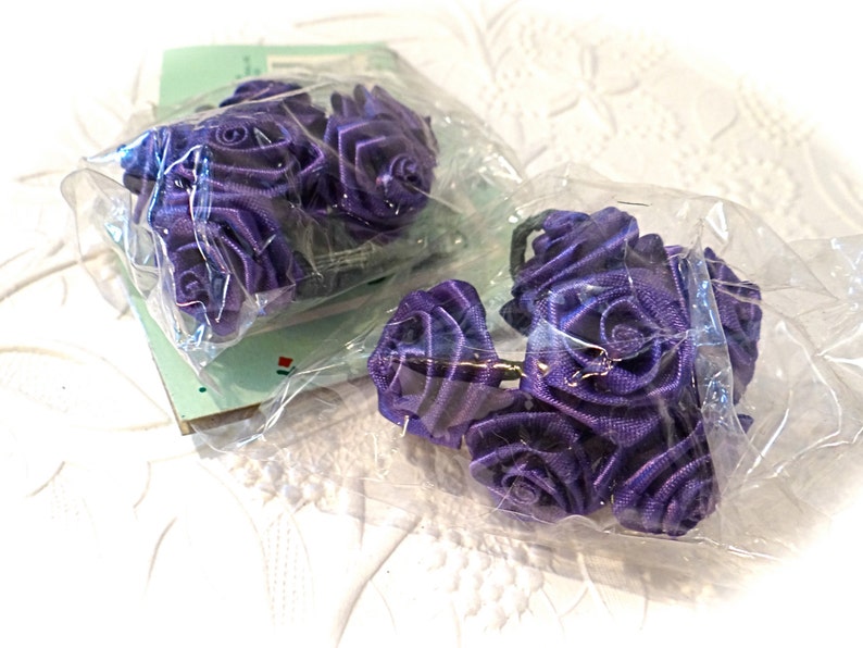 May include: Two packs of purple satin ribbon roses. The roses are arranged in a cluster and are wrapped in clear plastic.