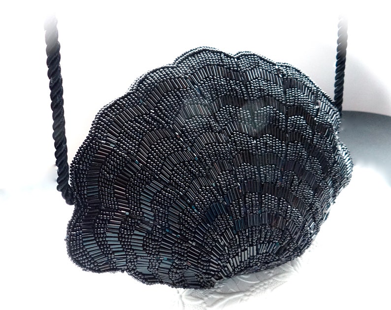 Puede incluir: Un bolso de noche negro de cuentas con forma de concha con una correa de cuerda negra.
