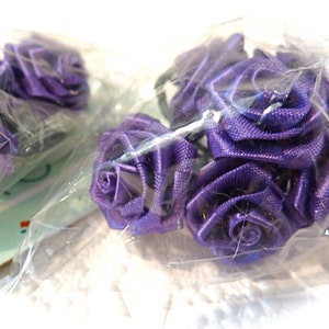 May include: A pack of purple satin ribbon roses. The roses are arranged in a cluster and are wrapped in clear plastic.