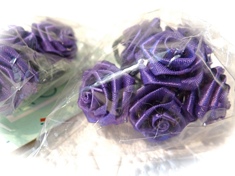 May include: A pack of purple satin ribbon roses. The roses are arranged in a cluster and are wrapped in clear plastic.
