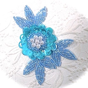 May include: A blue and white beaded flower applique with a center of white pearls. The flower has three leaves.