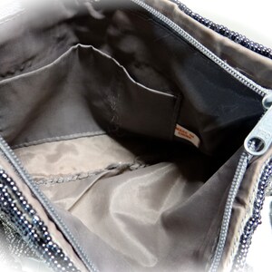May include: The inside of a black beaded handbag with a zip closure and a small pocket. The lining is a dark grey fabric. The tag reads "Made in China."