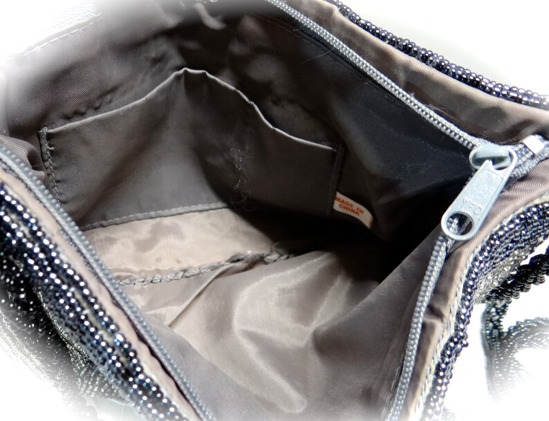 May include: The inside of a black beaded handbag with a zip closure and a small pocket. The lining is a dark grey fabric. The tag reads "Made in China."