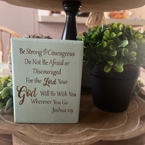 May include: A light green wooden block with a gold inscription that reads "Be Strong & Courageous Do Not Be Afraid or Discouraged For the Lord Your God Will Be With You Wherever You Go Joshua 1:9".