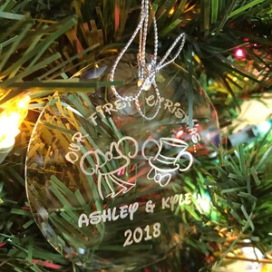 Pu&ograve; includere: Ornamento natalizio rotondo e trasparente con la scritta "Our First Christmas", i nomi "Ashley & Kyle" e l'anno "2018". L'ornamento, sospeso da un filo d'argento, presenta disegni di campana e cappello.