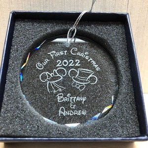 Pu&ograve; includere: Un ornamento natalizio rotondo e trasparente con testo e immagini incise, presentato in una scatola blu scuro. L'ornamento recita "Our First Christmas 2022" con i nomi "Brittany & Andrew" sotto. Ha un cordino d'argento per appenderlo.