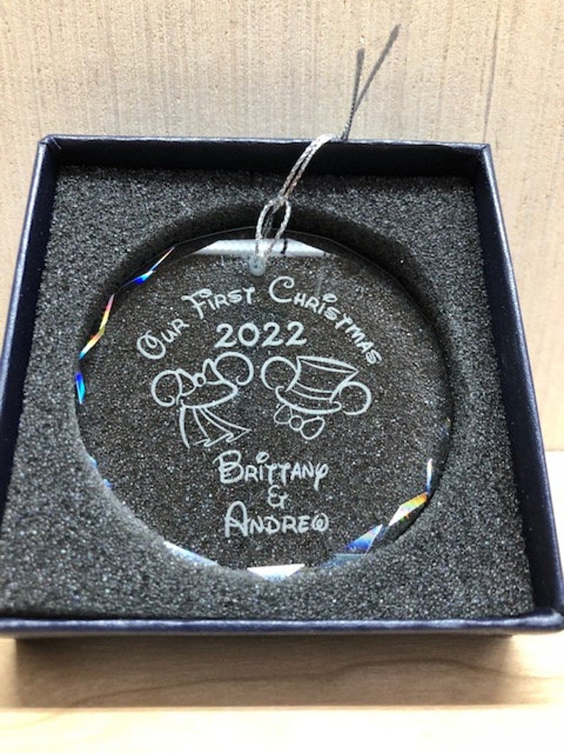 Pu&ograve; includere: Un ornamento natalizio rotondo e trasparente con testo e immagini incise, presentato in una scatola blu scuro. L'ornamento recita "Our First Christmas 2022" con i nomi "Brittany & Andrew" sotto. Ha un cordino d'argento per appenderlo.