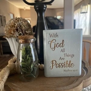 May include: A white wooden sign with the text "With God All Things Are Possible" in gold lettering and the Bible verse "Matthew 19:26" below.
