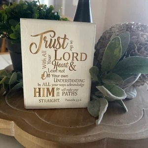 May include: White wooden sign with gold lettering that reads "Trust in the LORD with all your Heart & lean not on your own understanding. In ALL your ways acknowledge HIM, and He will make your paths STRAIGHT. Proverbs 3:5-6"