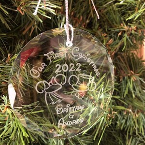 Pu&ograve; includere: Una palla di Natale rotonda e trasparente con la scritta "Our First Christmas 2022" e i nomi "Brittany & Andreo". La palla &egrave; appesa a un albero di Natale.