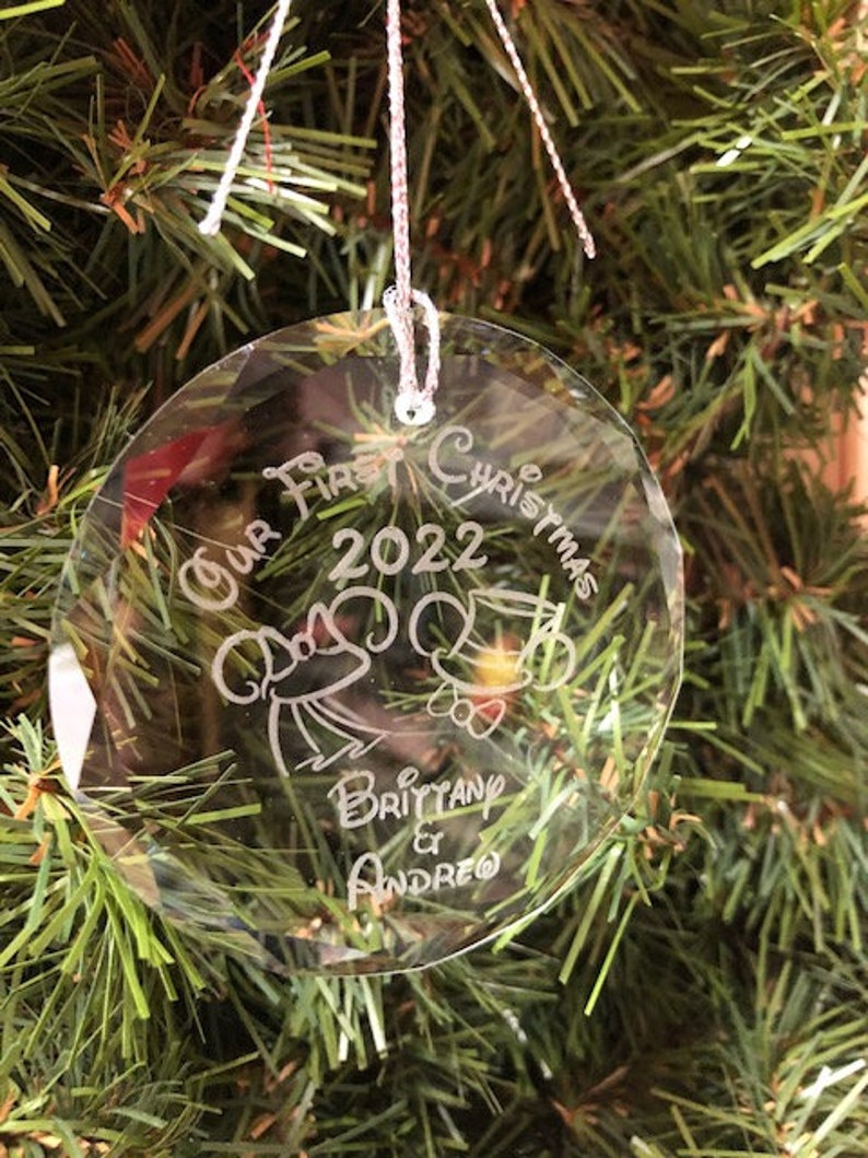Pu&ograve; includere: Una palla di Natale rotonda e trasparente con la scritta "Our First Christmas 2022" e i nomi "Brittany & Andreo". La palla &egrave; appesa a un albero di Natale.