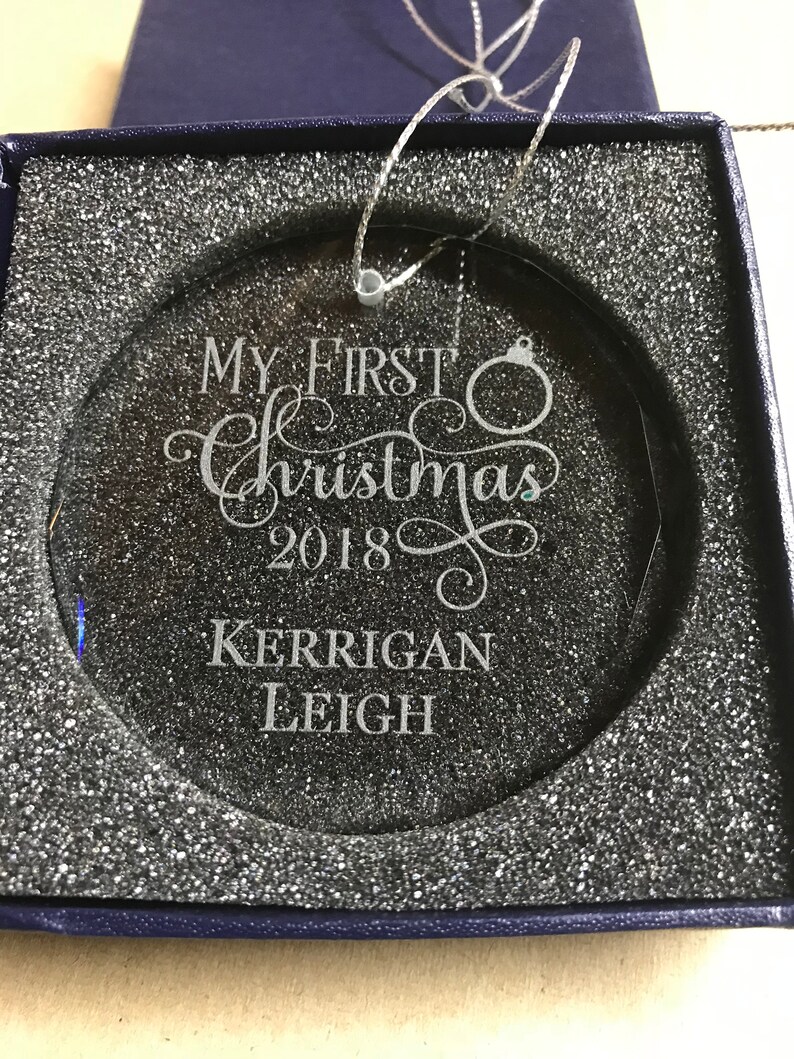 May include: A clear glass Christmas ornament with the text "My First Christmas 2018" and the name "Kerrigan Leigh" engraved on it. The ornament is in a blue box with white foam padding.