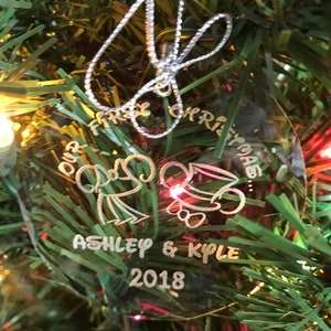 Pu&ograve; includere: Ornamento natalizio rotondo e trasparente con le parole "Our First Christmas", i nomi "Ashley & Kyle" e l'anno "2018". L'ornamento &egrave; appeso a un albero di Natale.