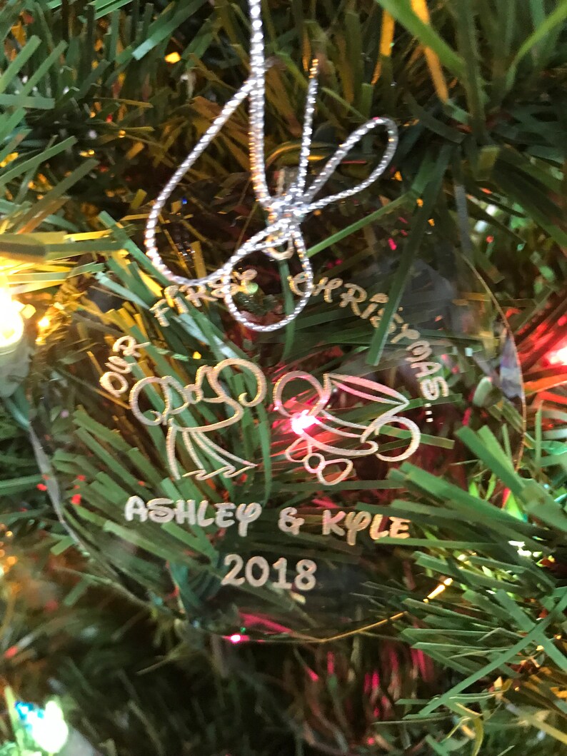 Pu&ograve; includere: Ornamento natalizio rotondo e trasparente con le parole "Our First Christmas", i nomi "Ashley & Kyle" e l'anno "2018". L'ornamento &egrave; appeso a un albero di Natale.