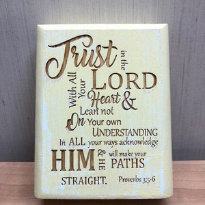 May include: A light yellow wooden sign with a white background and brown lettering. The sign reads "Trust in the Lord with all your Heart & Lean not on your own Understanding. In all your ways acknowledge Him & He will make your paths straight. Proverbs 3:5-6"