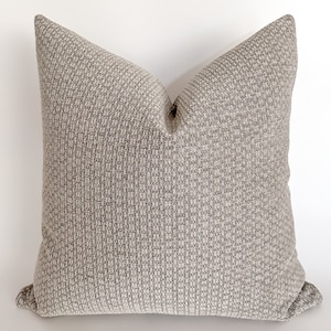 May include: A square throw pillow with a textured, woven fabric in a light gray color. The pillow has a subtle pattern of small squares.