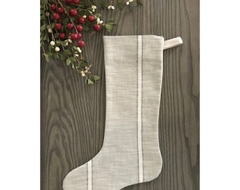 May include: A gray and white striped Christmas stocking with a white loop at the top. The stocking is on a wood background.
