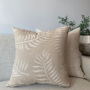 May include: Two square beige throw pillows with a white leaf pattern. One pillow is in the foreground, displaying the leaf design, while the other is in the background, with a solid beige texture. The pillows are on a light gray sofa.