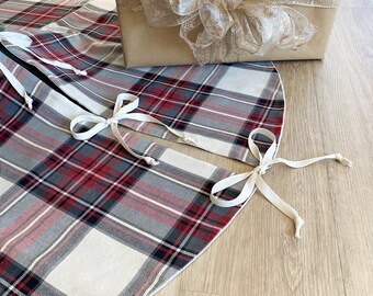 May include: A white and red plaid Christmas tree skirt with white ribbon ties. The skirt is on a wood floor and a wrapped gift box is in the background.