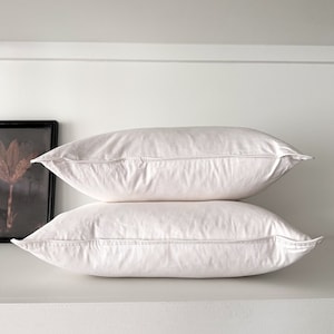 May include: Two white pillowcases stacked on top of each other on a white shelf. The pillowcases are made of a soft, smooth fabric.