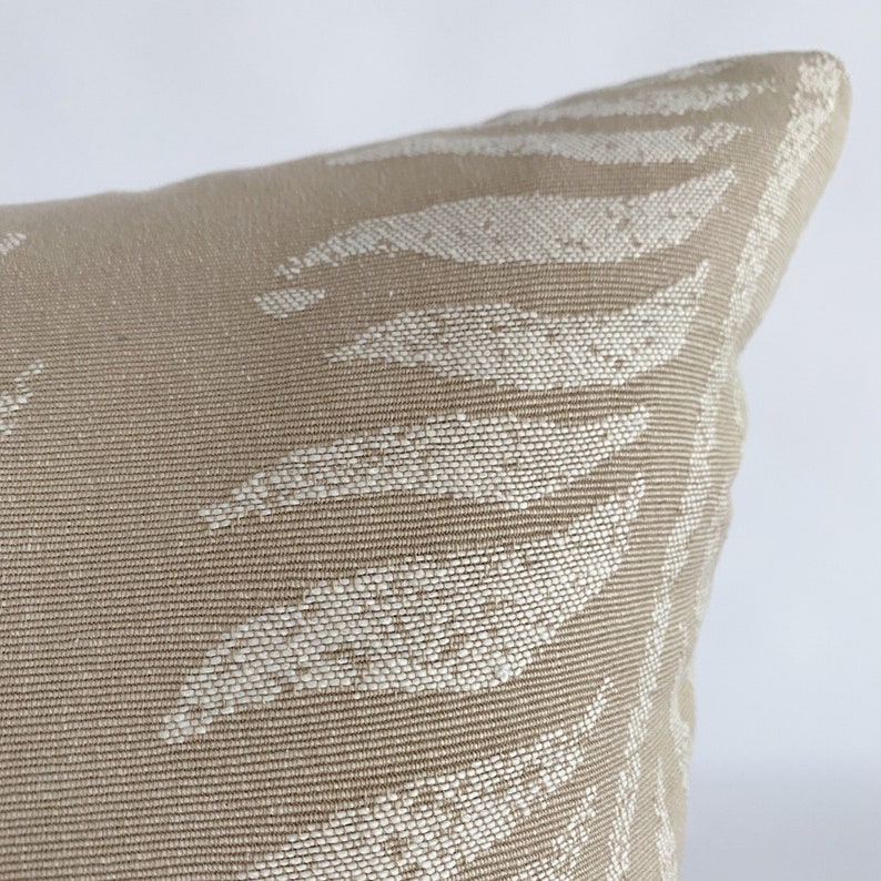 May include: Close-up of a beige decorative pillow with a woven texture. The pillow features a repeating pattern of white leaf-like shapes. The pillow has a soft, inviting appearance, suitable for home decor.