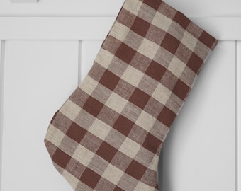 May include: A brown and beige checkered Christmas stocking hanging from a wooden peg. The stocking is made of a woven fabric with a classic buffalo plaid pattern. The background is a white wall.