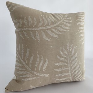 May include: A square beige throw pillow with a repeating pattern of white fern leaves. The pillow has a textured appearance and is likely made of fabric. The design is simple and would complement a variety of home decor styles.