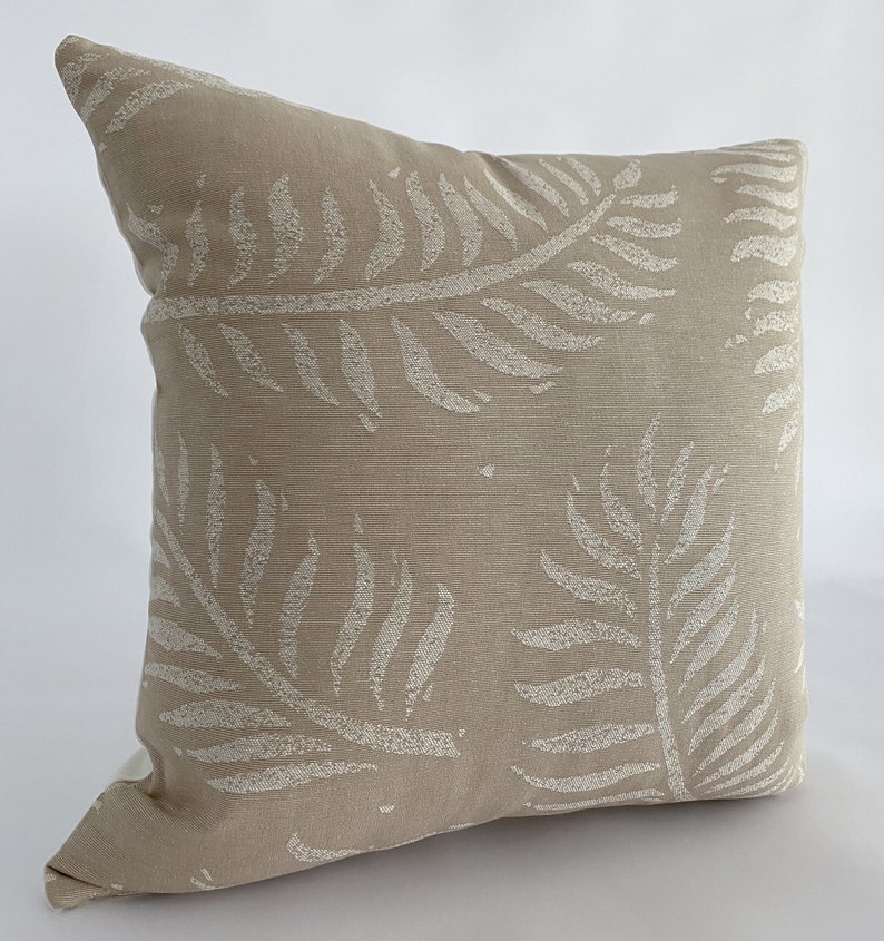 May include: A square beige throw pillow with a repeating pattern of white fern leaves. The pillow has a textured appearance and is likely made of fabric. The design is simple and would complement a variety of home decor styles.