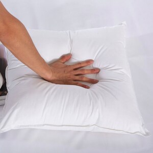 May include: A white square pillow with a hand pressing down on the centre of the pillow.
